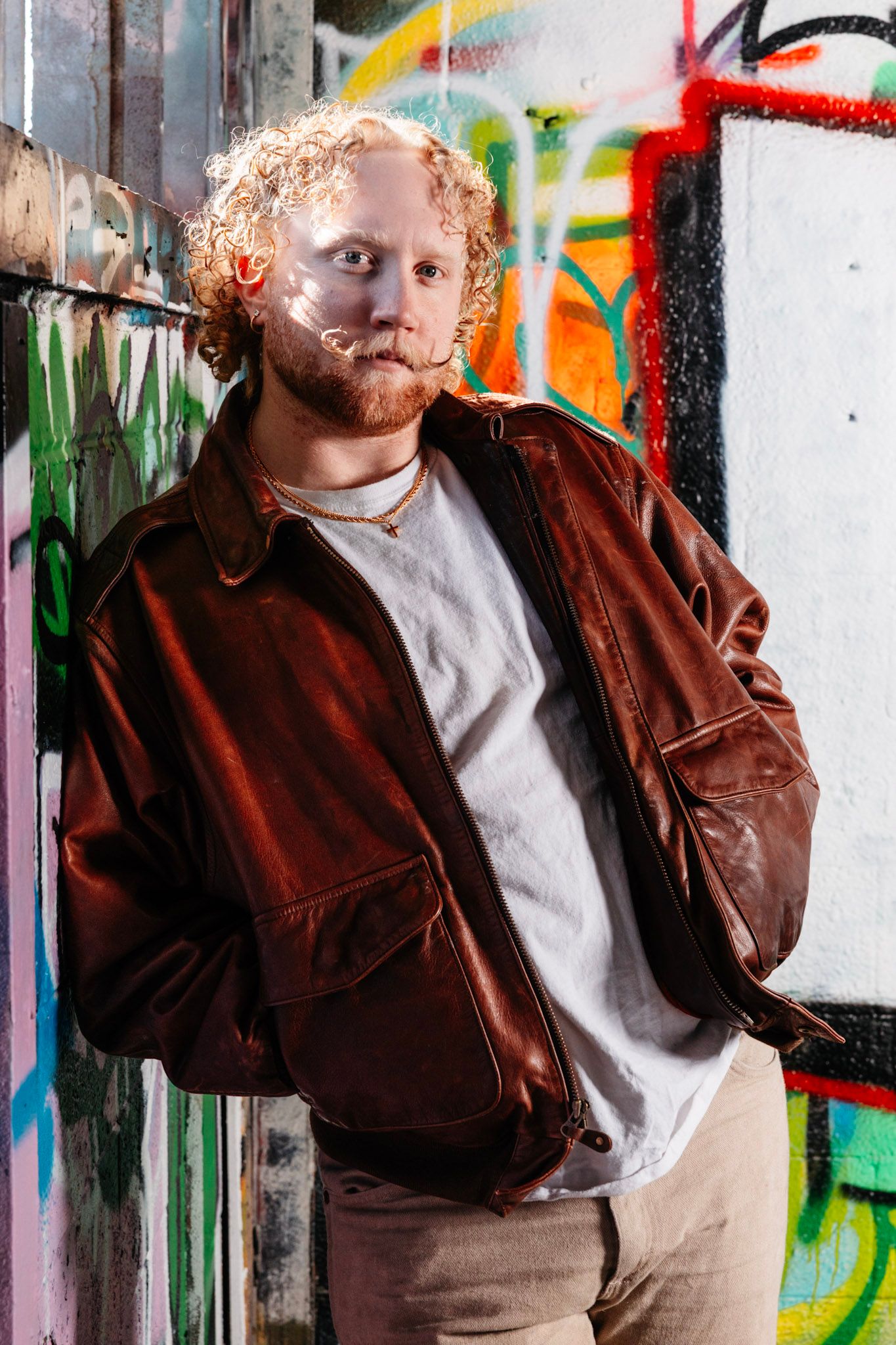 A person in a brown leather jacket poses against a graffiti wall with colorful street art in the background.