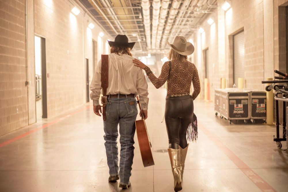 Two performers in western attire walk down a backstage hallway while holding hands and carrying a guitar case.