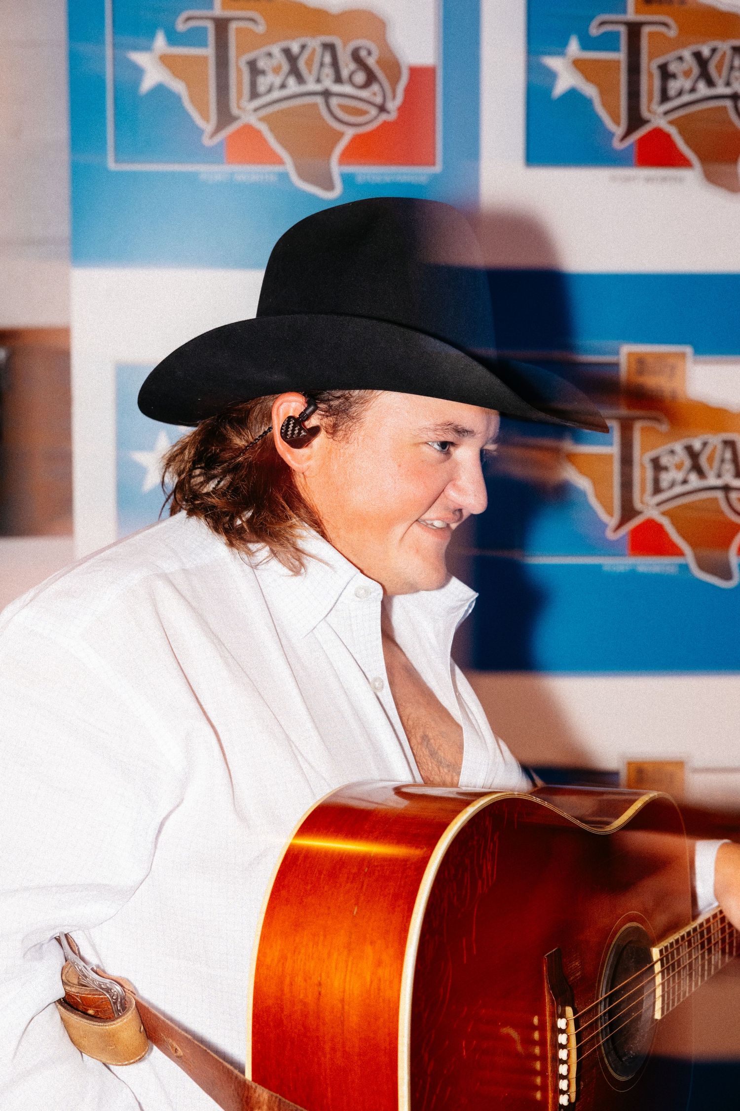 A musician in a white shirt and cowboy hat plays an acoustic guitar against a blue background with Texas logos.