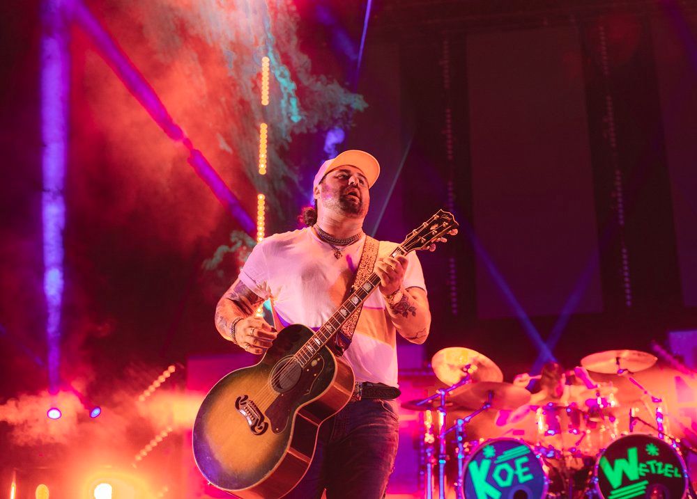 Musician Koe Wetzel plays acoustic guitar on stage with dramatic red and purple concert lighting.