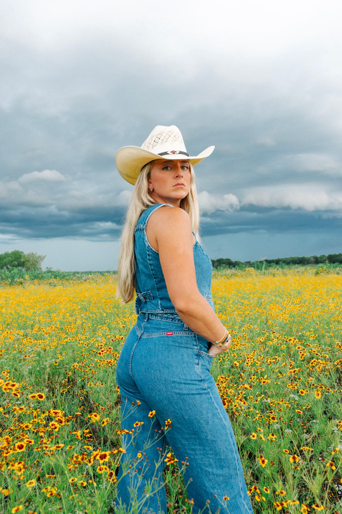 Woman in denim overalls and white cowboy hat standing in yellow wildflower field under stormy sky.