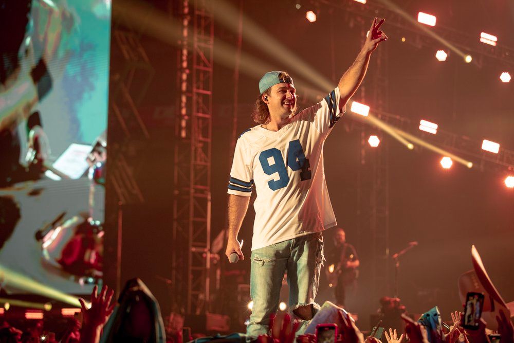 Concert performer Morgan Wallen in a Dallas sports jersey energetically entertaining crowd with dramatic lighting.