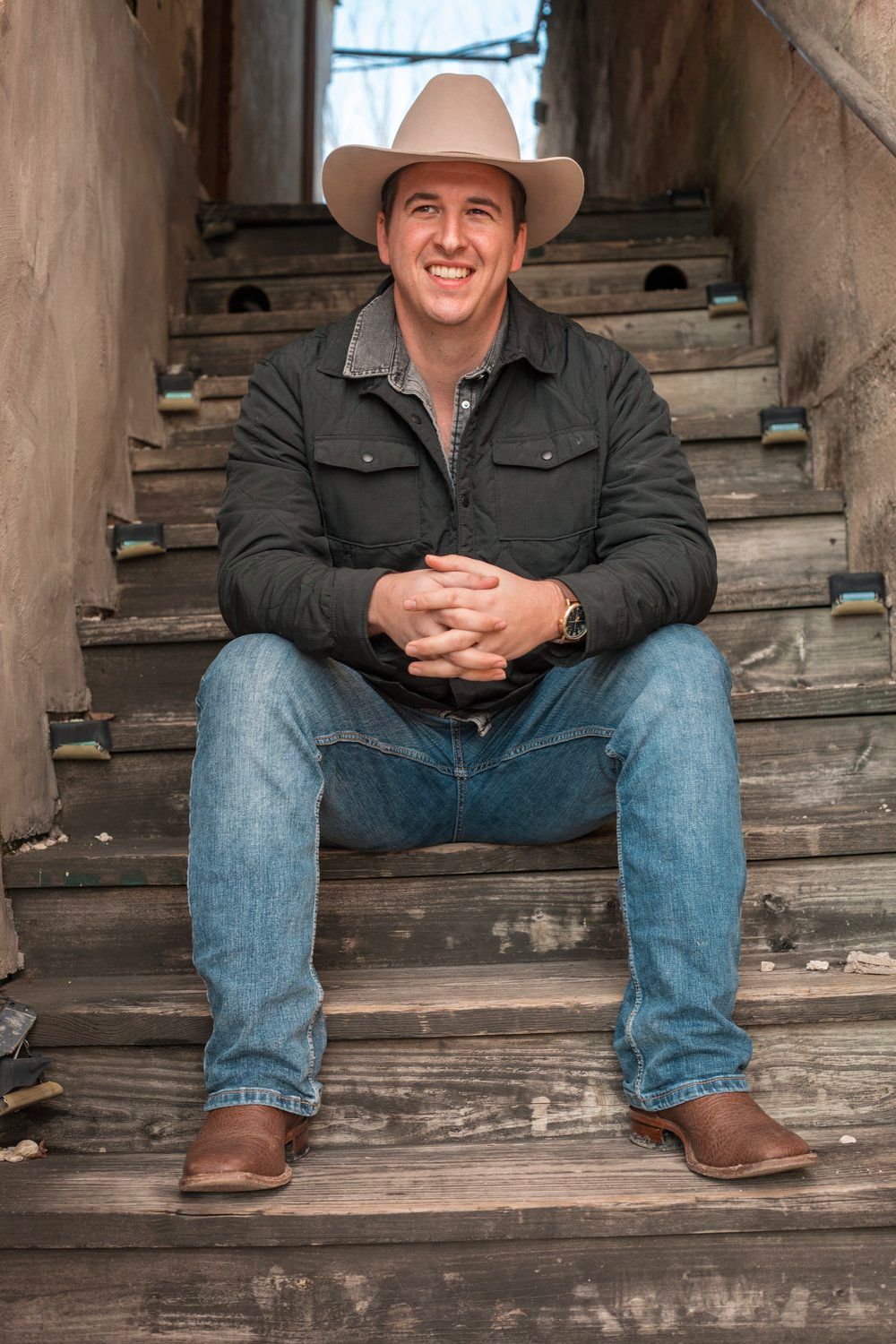 Western style portrait of someone seated on rustic wooden stairs wearing cowboy hat and denim.