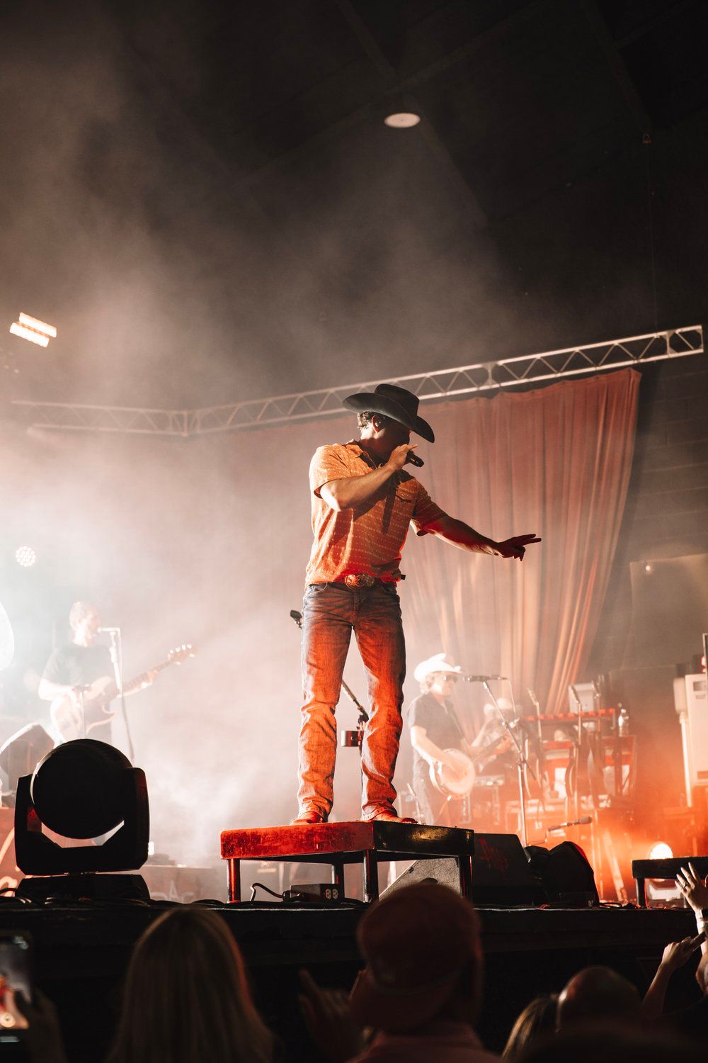 Concert performer in orange outfit stands on platform amid dramatic stage lighting and fog effects.