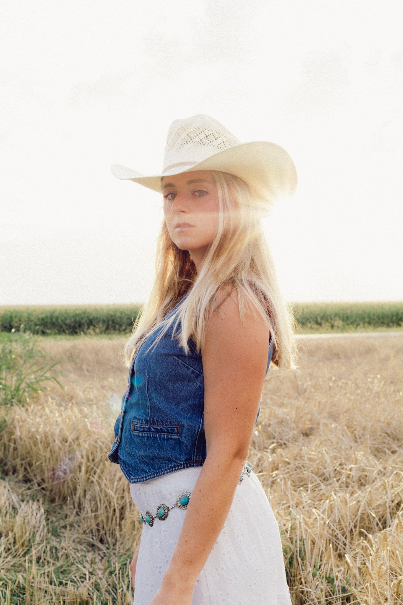 Blonde woman in cowboy hat, denim vest, and white skirt stands in golden field with bright backlight.