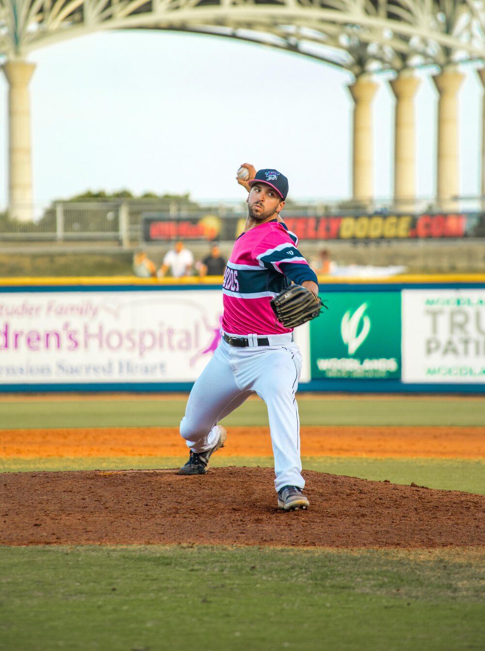 Baseball pitcher in white and pink uniform winds up to throw from the mound during a minor league game.