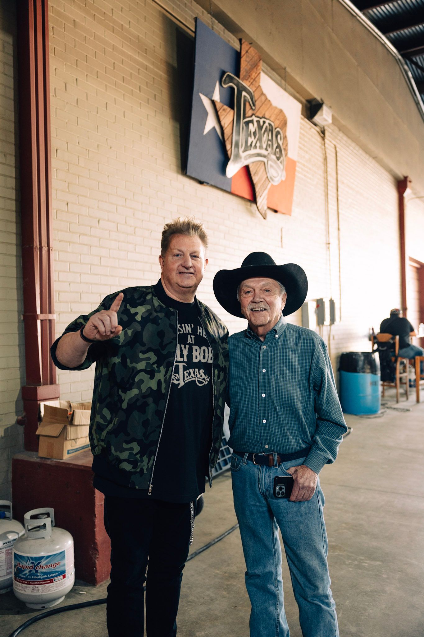 Two men posing indoors under a Texas sign, one in camo jacket giving thumbs up, other in cowboy hat and blue shirt.