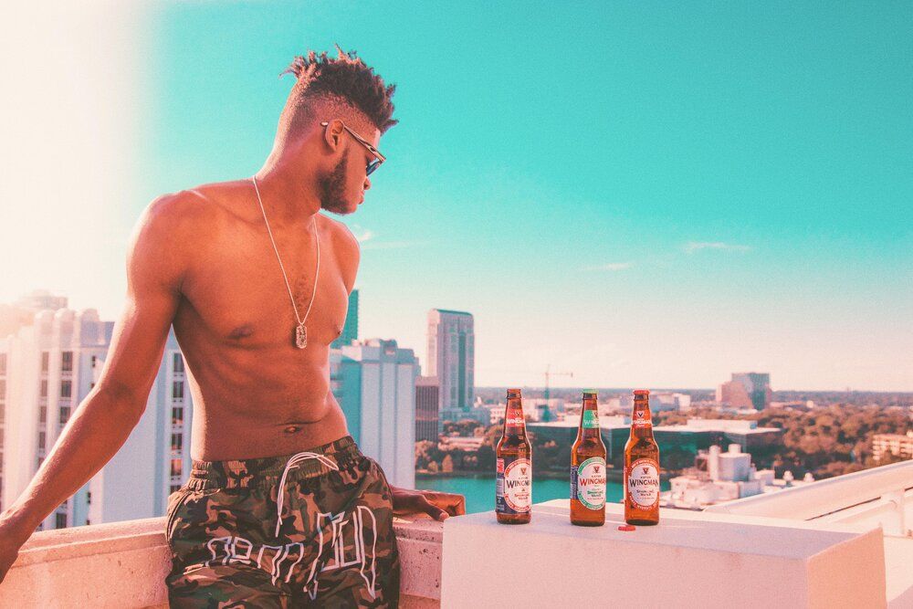 Person standing shirtless on urban rooftop with beverages and city skyline in background against bright blue sky.