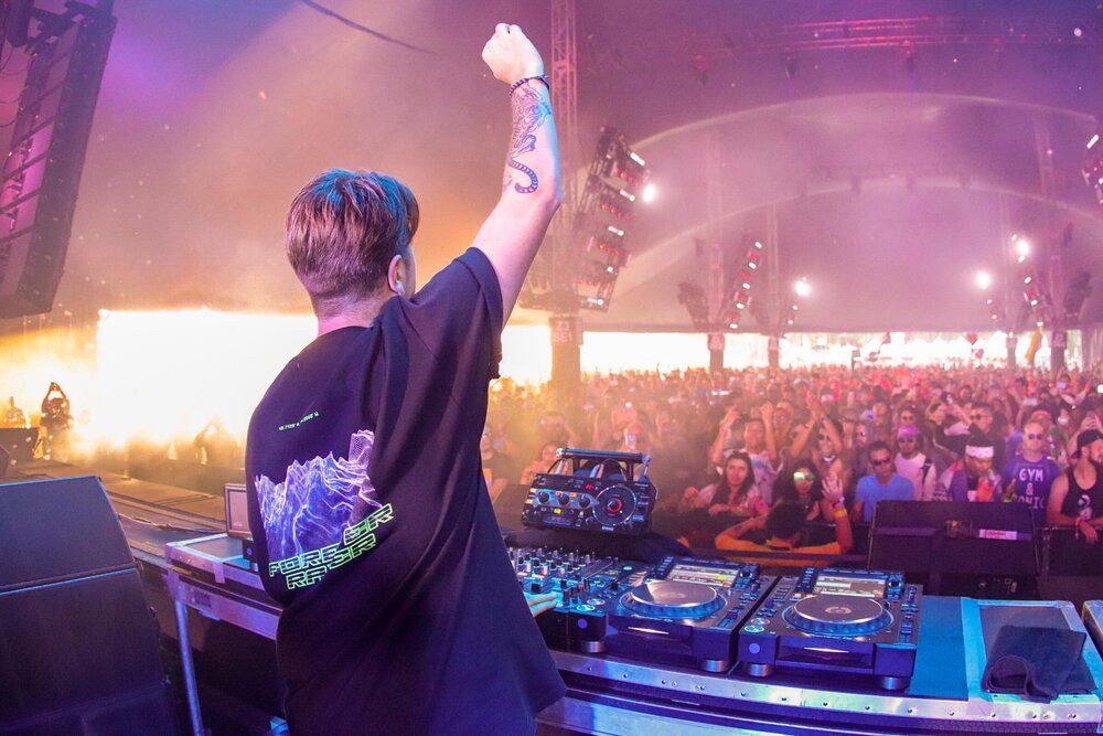 DJ raises hand to crowd while performing at electronic music festival with colorful ambient lighting.