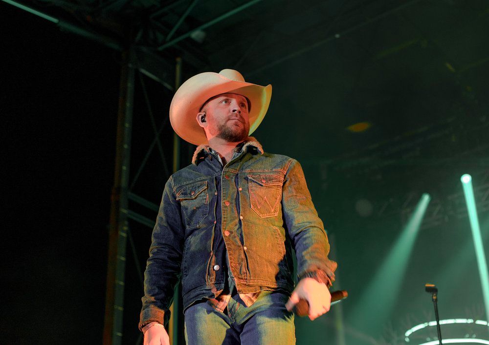 Country performer  Justin Moore in denim jacket and cowboy hat sings on stage with green lighting.