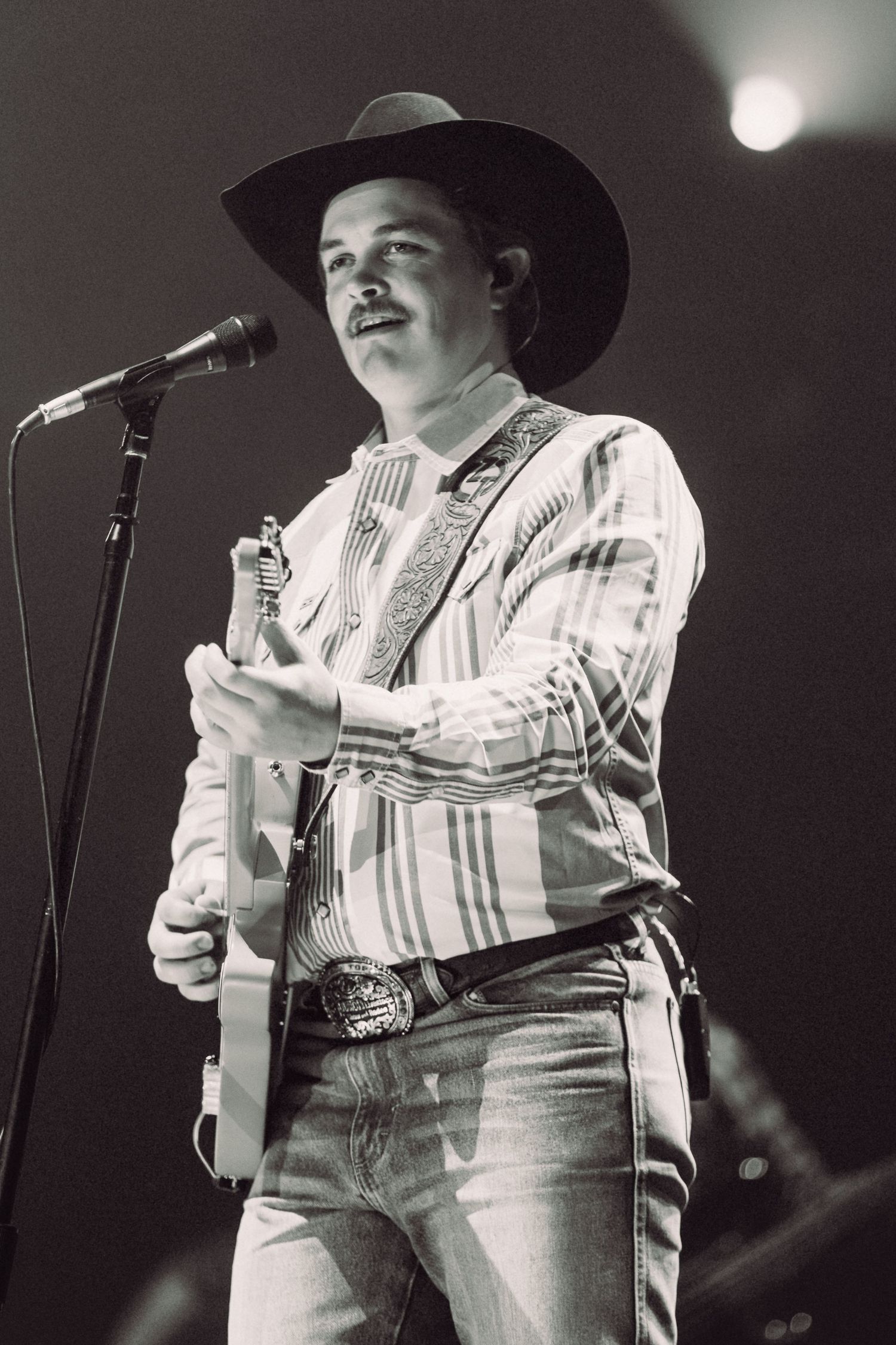Country music performer in cowboy hat and striped shirt sings into microphone during concert performance.