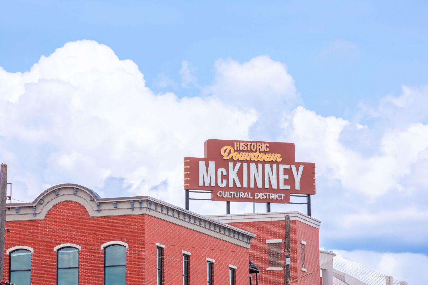 Historic Downtown McKinney Cultural District sign on a red brick building under a blue sky with white clouds.