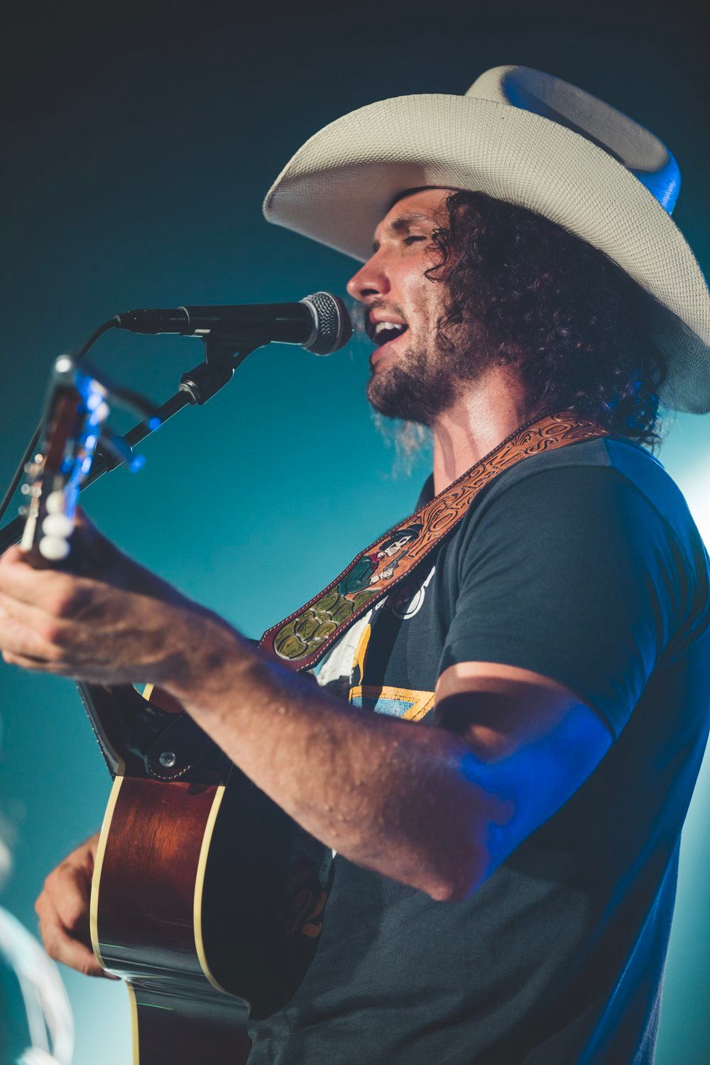 Country musician Josh Meloy in cowboy hat performs with acoustic guitar under blue stage lighting.
