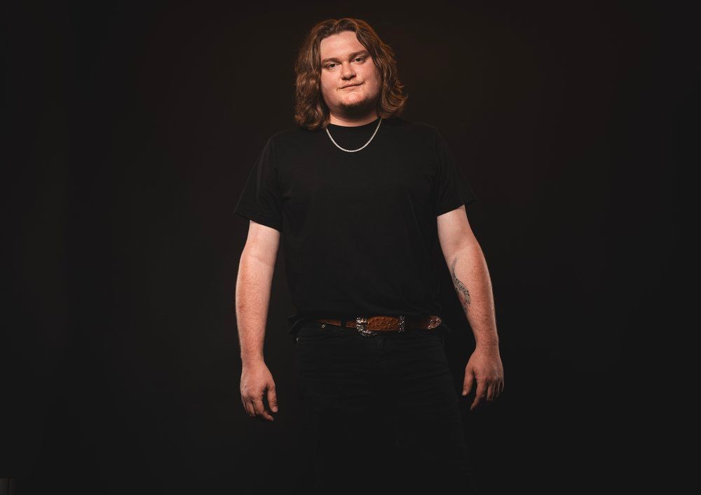 Person with long hair poses dramatically against black background wearing all black attire.