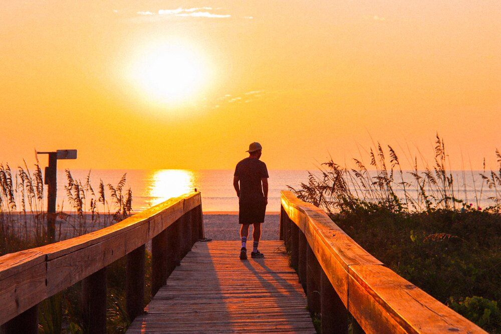 A silhouette stands on a wooden boardwalk path at sunset with tall grass and ocean views.