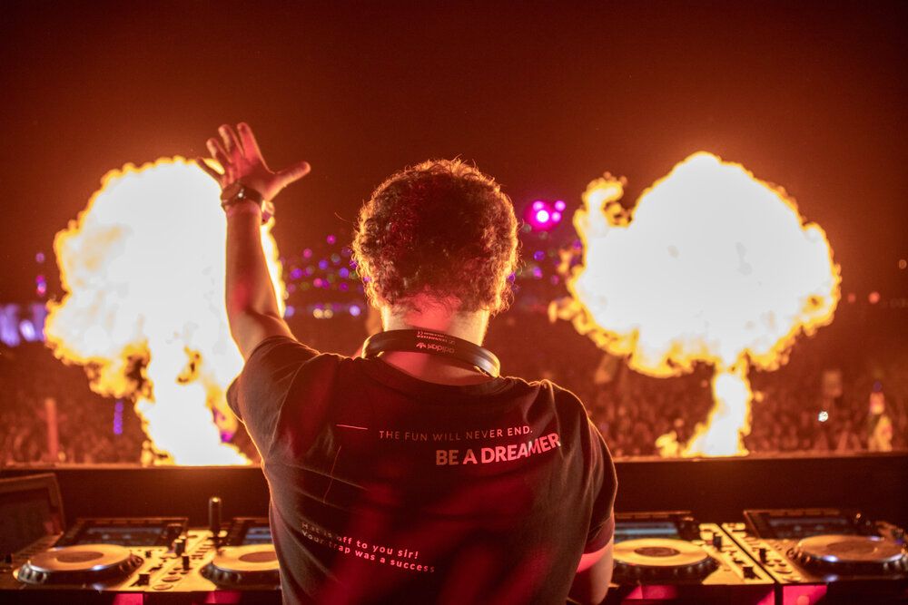 DJ performs on stage with dramatic fire effects and pyrotechnics bursting behind the decks at a music festival.