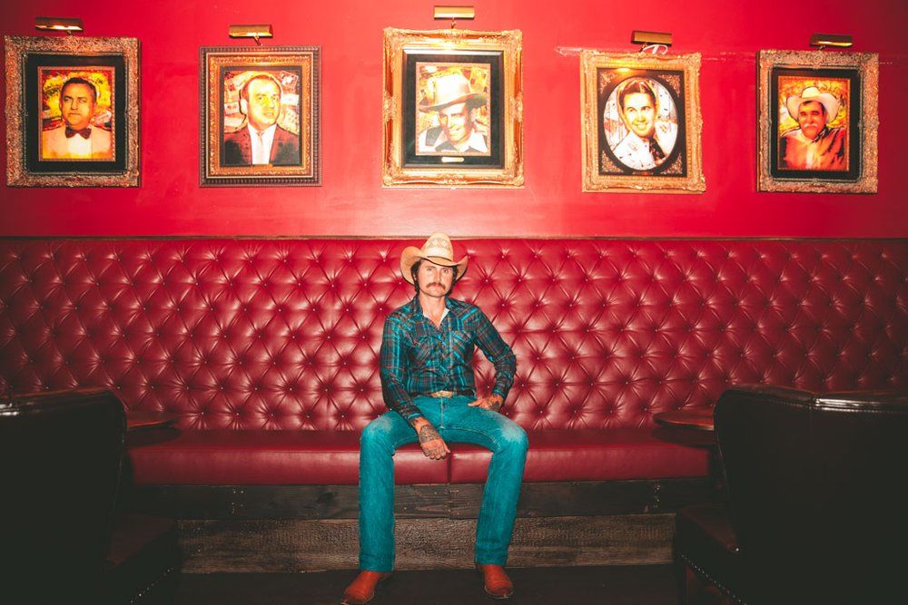 Musician in denim outfit sits on red leather booth beneath vintage framed portraits.