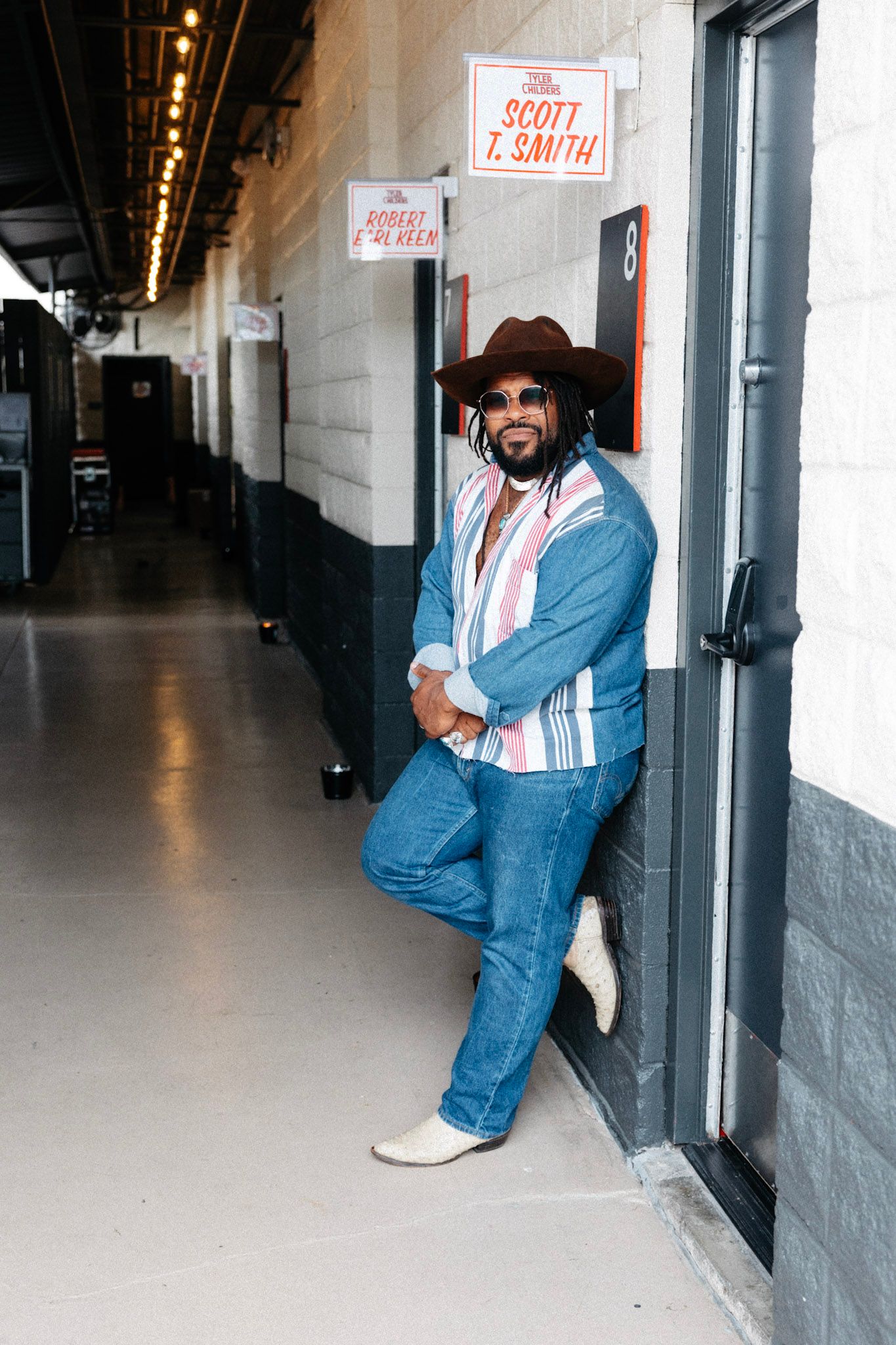 Man in cowboy hat, sunglasses, and jeans leans against wall in hallway near a door labeled Scott T. Smith.