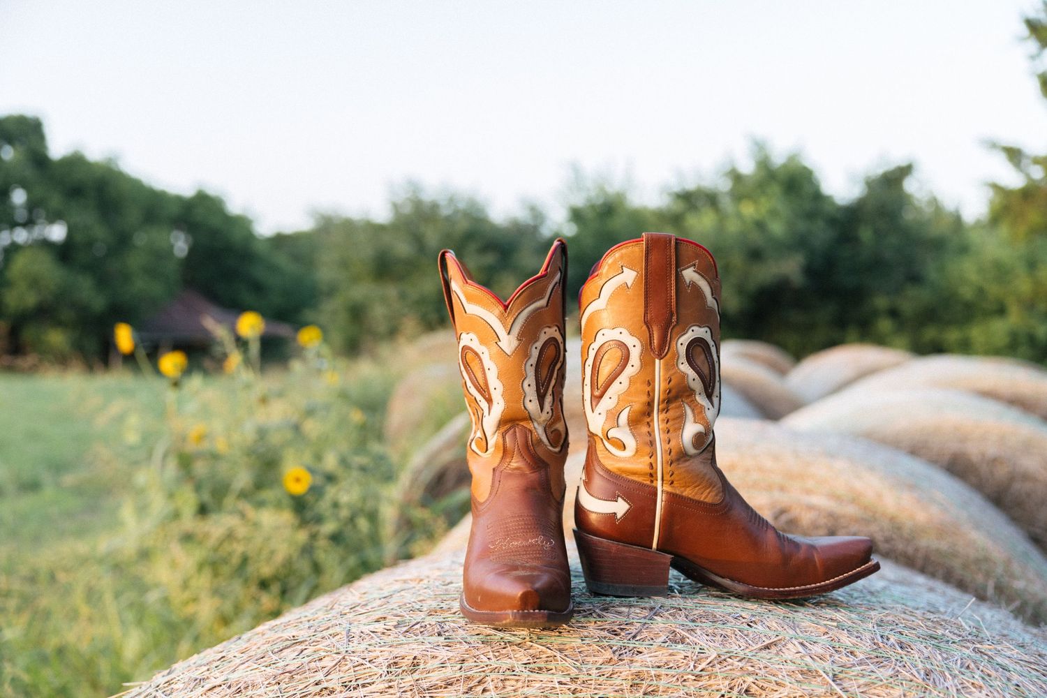 Brown leather cowboy boots with white stitching resting on a hay bale in a sunny rural field.