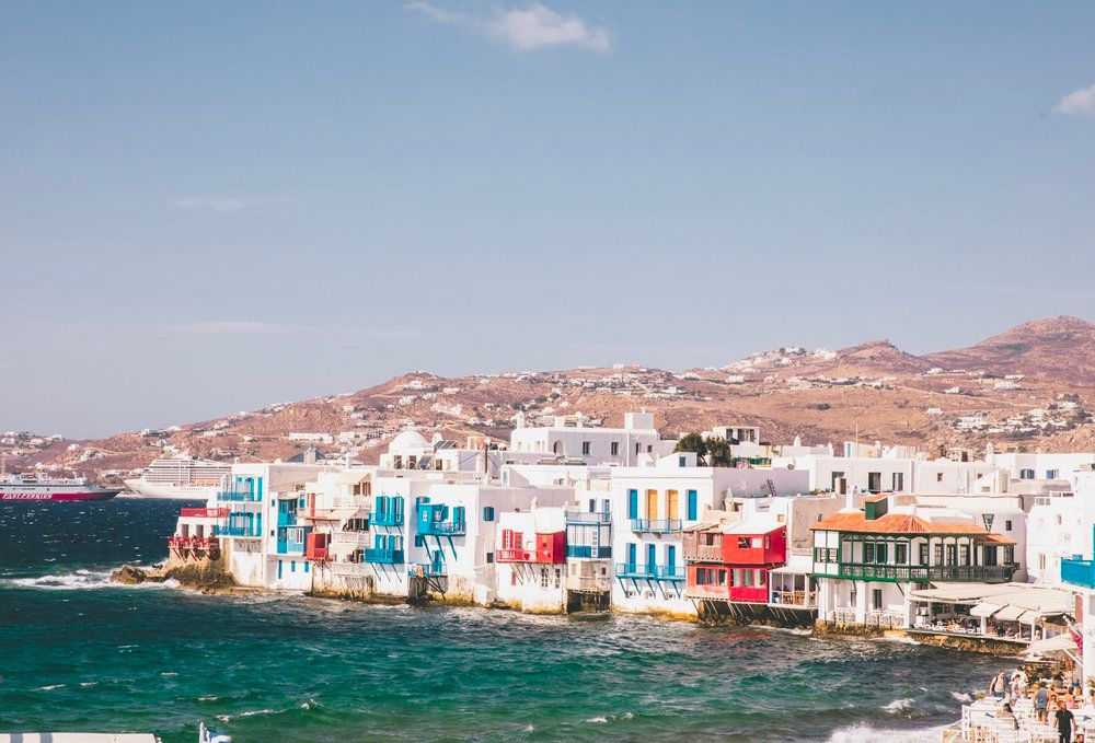 Colorful waterfront buildings line the coast of Mykonos, Greece against turquoise Mediterranean waters.