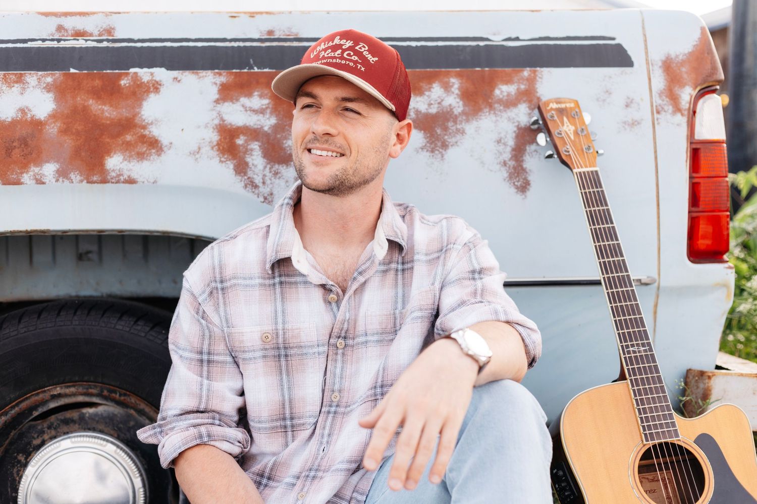 Man in plaid shirt and red cap sits by vintage truck with acoustic guitar, relaxed outdoor country music artist photo.