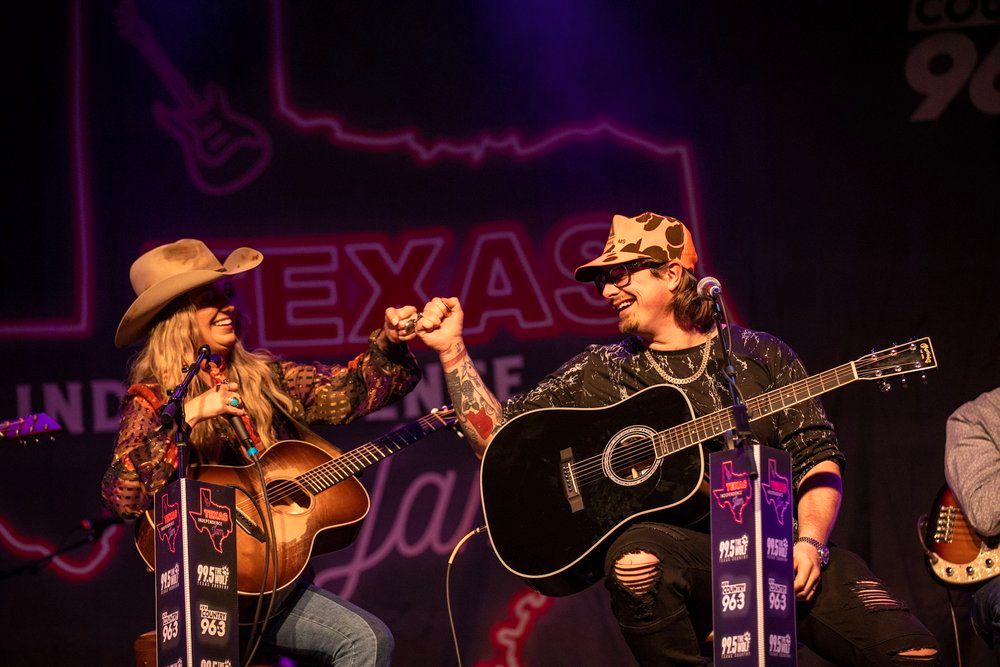 Country music performers with cowboy hats Hardy & Lainey Wilson play acoustic guitars together on a dimly lit stage.
