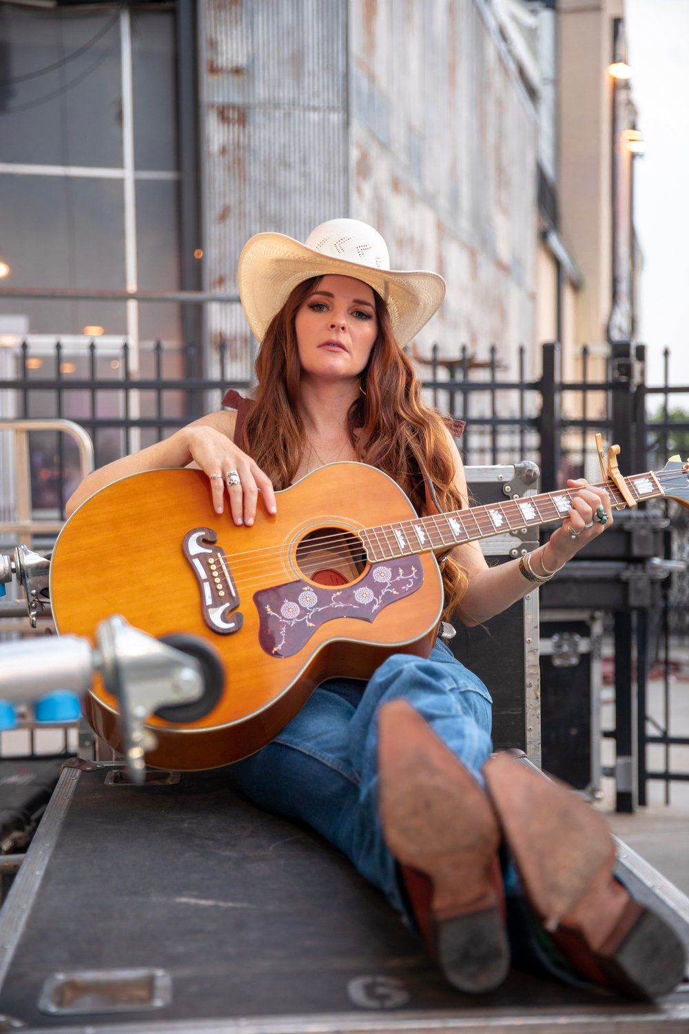 Country musician in cowboy hat playing acoustic guitar while sitting on equipment case.