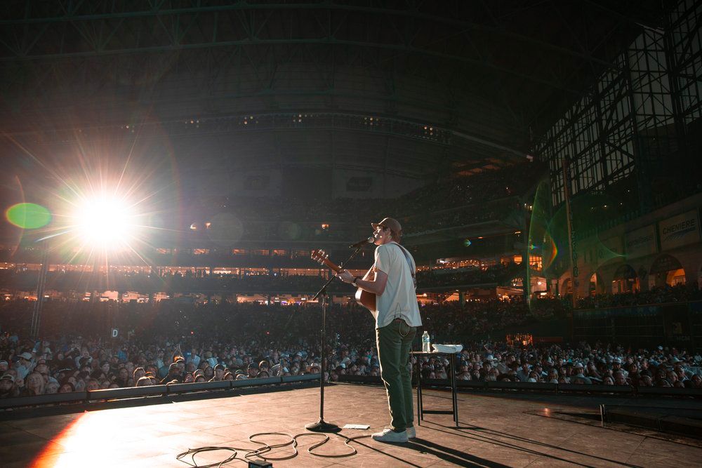 Performer Dylan Gossett on stage in front of massive stadium crowd with bright spotlight illumination.