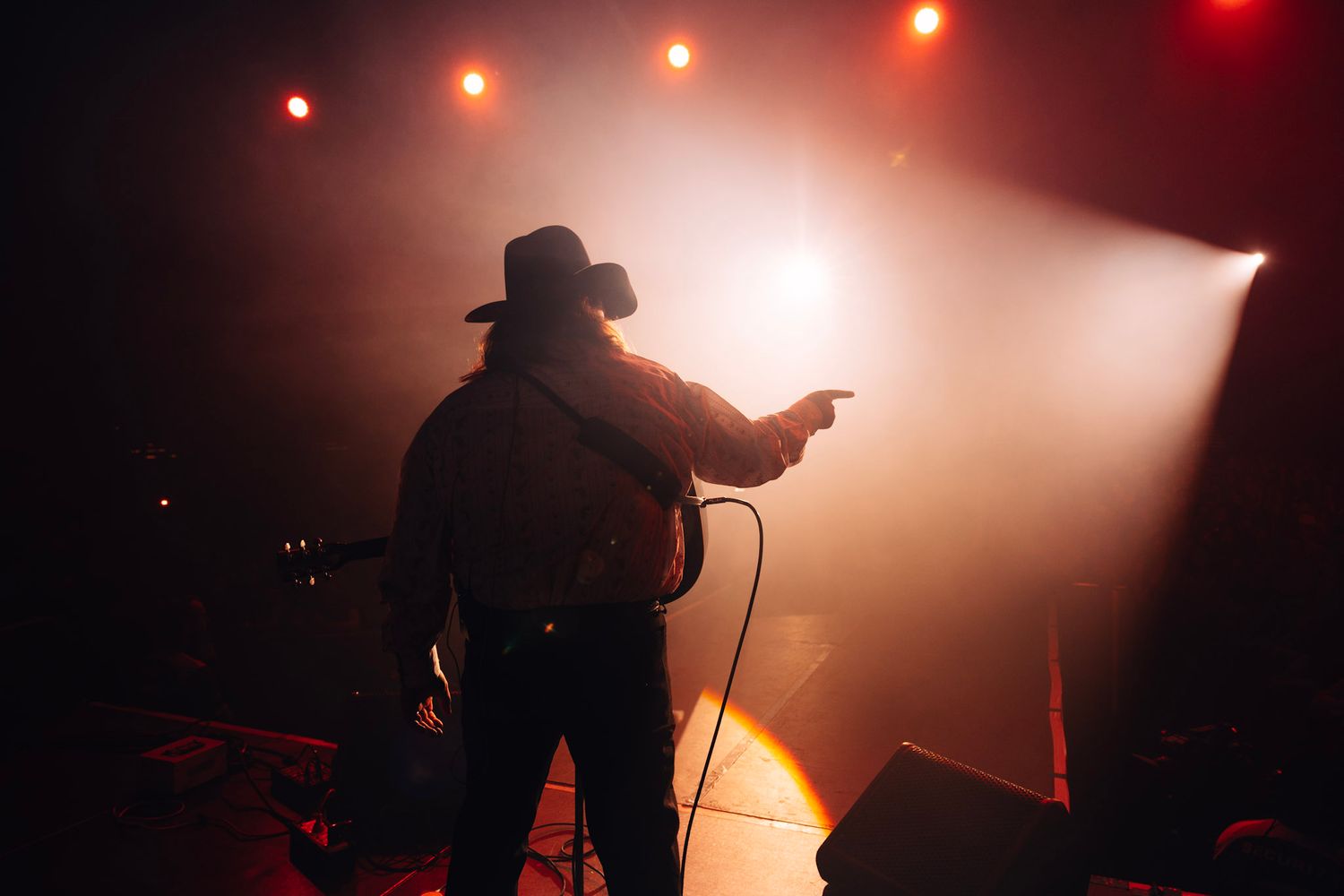 Silhouetted country musician in cowboy hat performing on stage with dramatic red and orange spotlights.