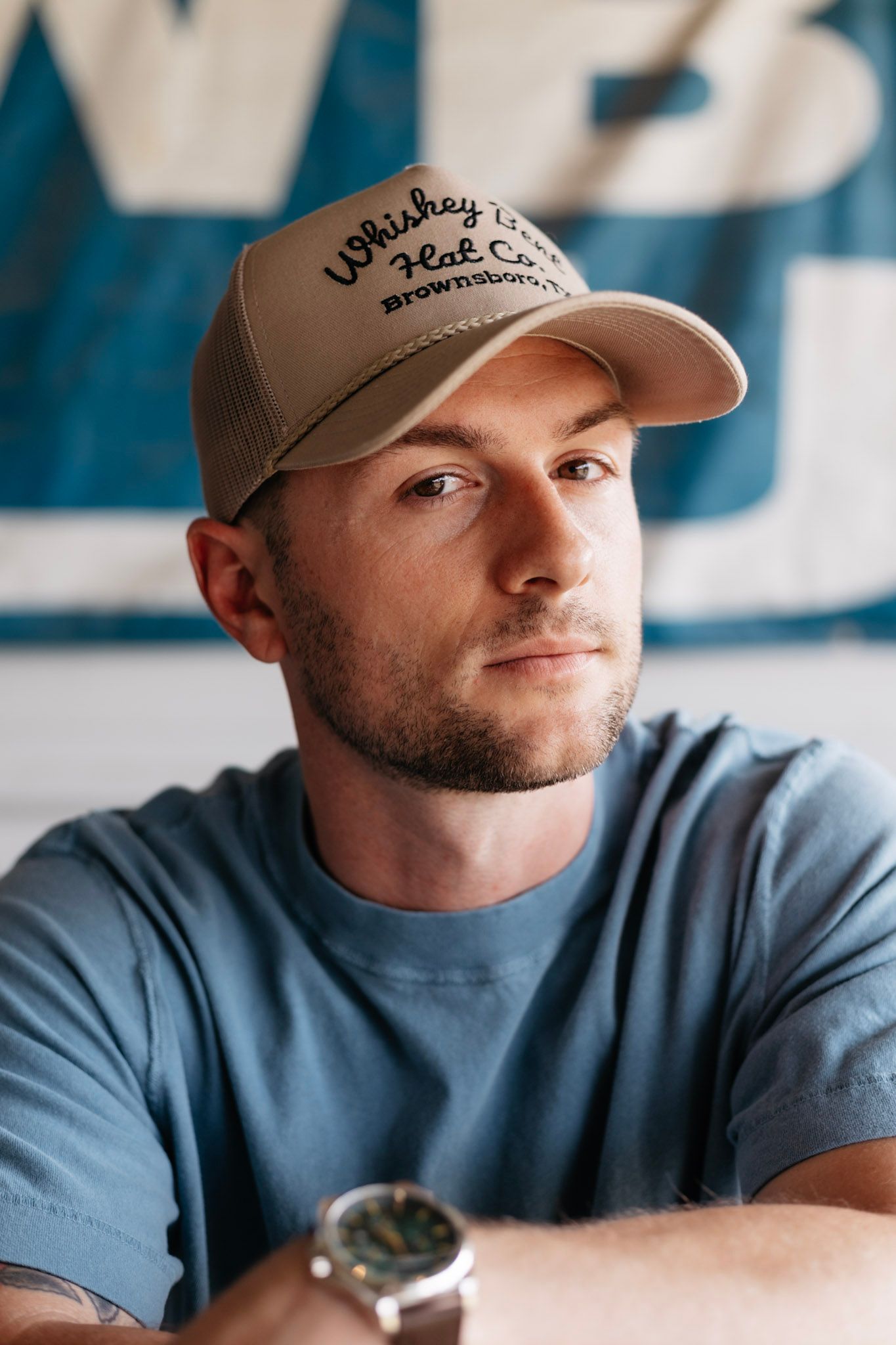 A person in a blue t-shirt and baseball cap sits casually against a blue background wearing a wristwatch.
