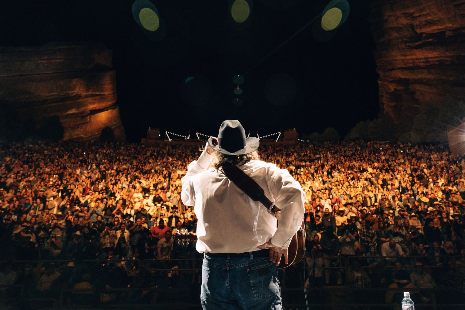 Silhouetted performer on stage in front of massive illuminated crowd at outdoor concert venue.