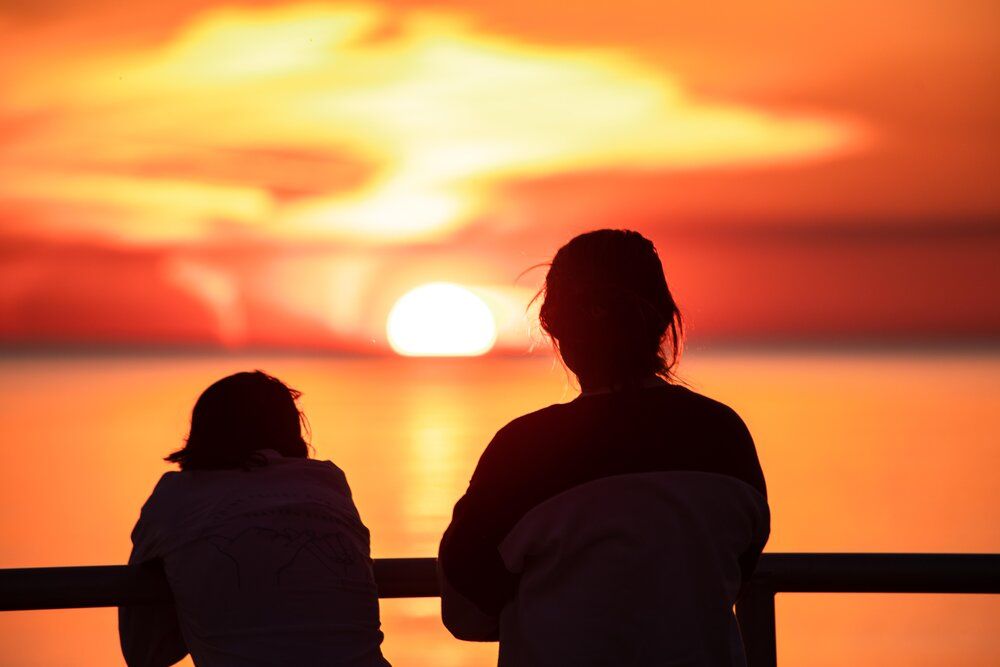 Silhouettes of people watching a vibrant orange sunset over the water with clouds in the sky.