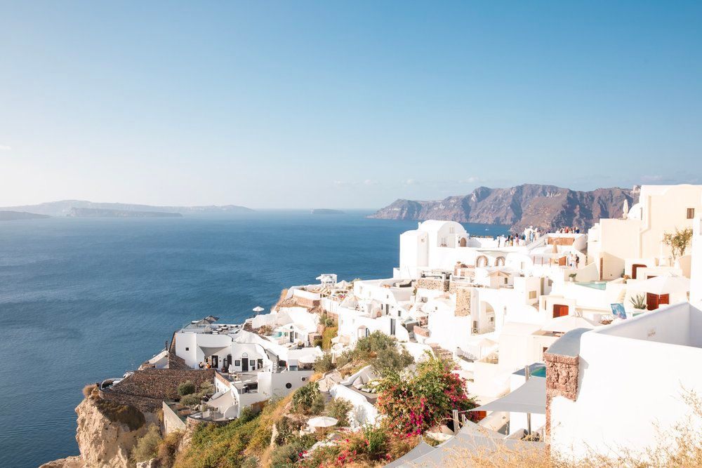 Aerial view of white buildings cascading down cliffs in Santorini Greece with blue Mediterranean sea and mountains.