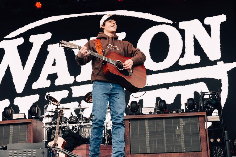 A performer stands on stage with a guitar in front of large white block letters during an outdoor concert performance.