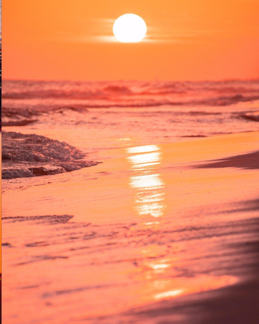 Dramatic orange sunset over ocean waves crashing on beach with sun reflection glowing on wet sand.