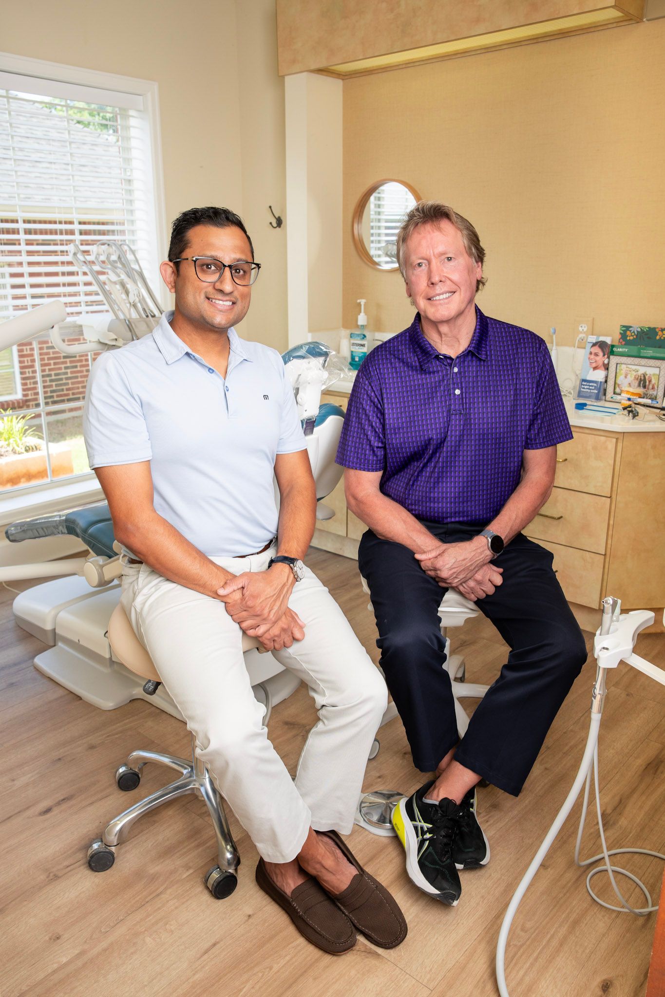 Two men smile together in a dental office, one in a light blue shirt and one in a purple polo shirt.