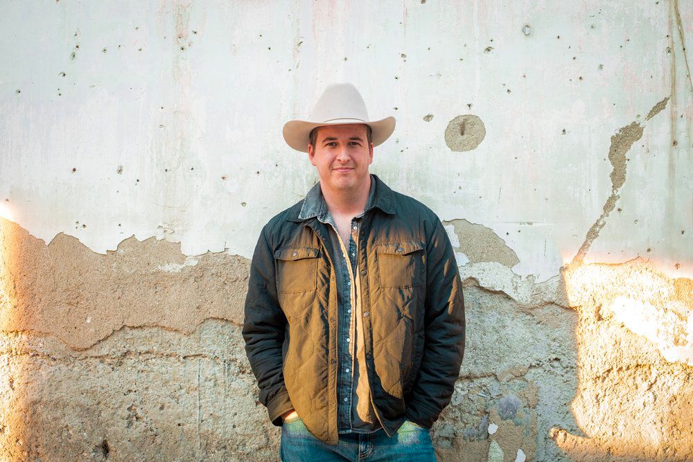 Country western style portrait against weathered wall wearing cowboy hat and jacket.