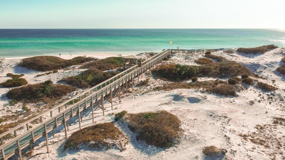 Wooden boardwalk winding through white sand dunes towards turquoise ocean waters on a sunny beach day.