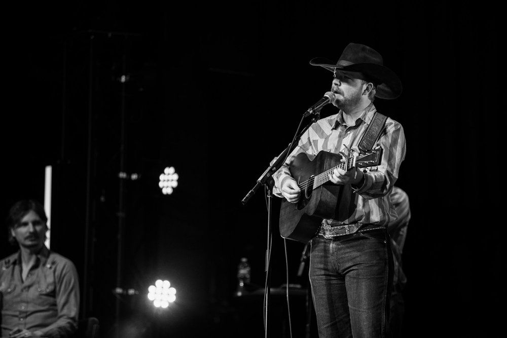 Country musician Colter Wall performs on stage in black and white wearing a cowboy hat and denim while playing acoustic guitar.