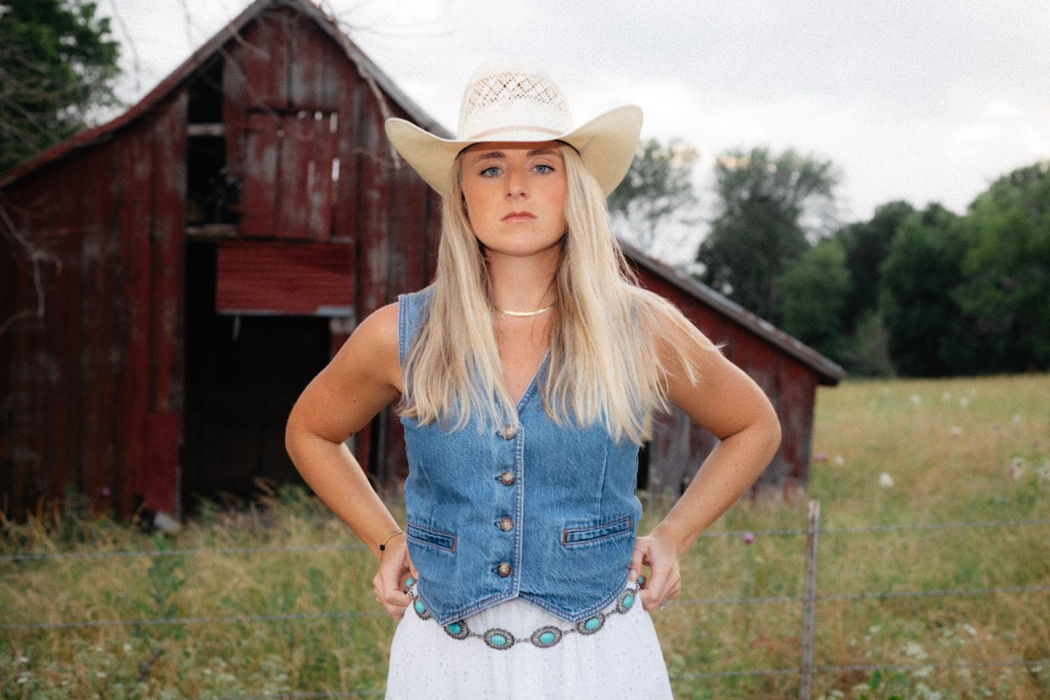 Blonde woman in cowboy hat and denim vest stands confidently in a field with a red barn behind her.