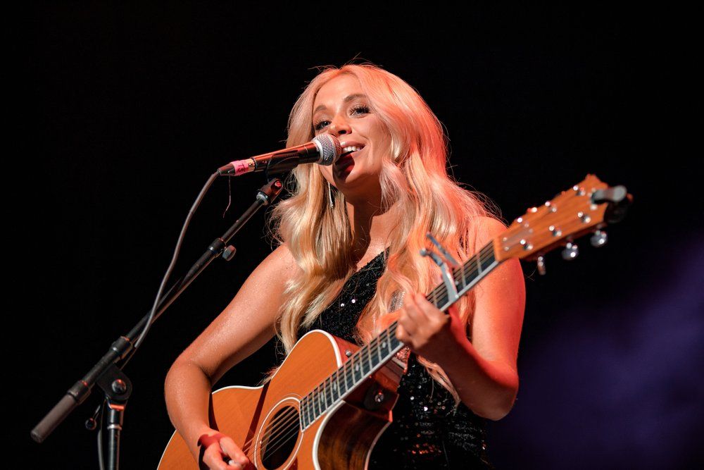 Female country singer Megan Moroney performs with acoustic guitar under purple stage lighting.