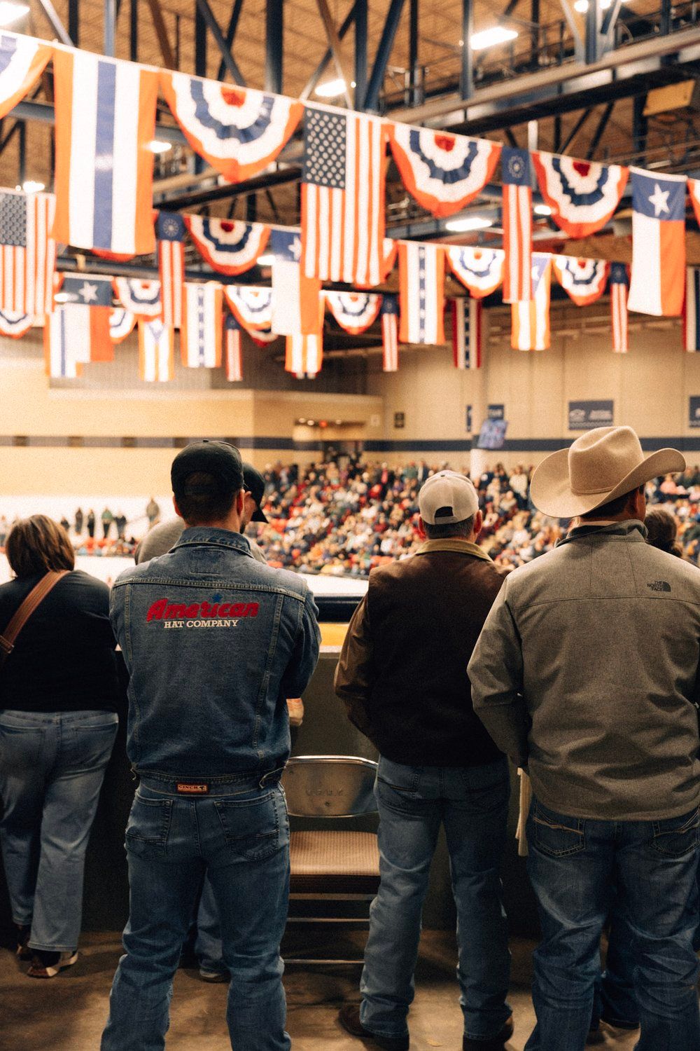 Spectators stand in blue jeans watching an indoor event with American flags and patriotic bunting decorating the ceiling.