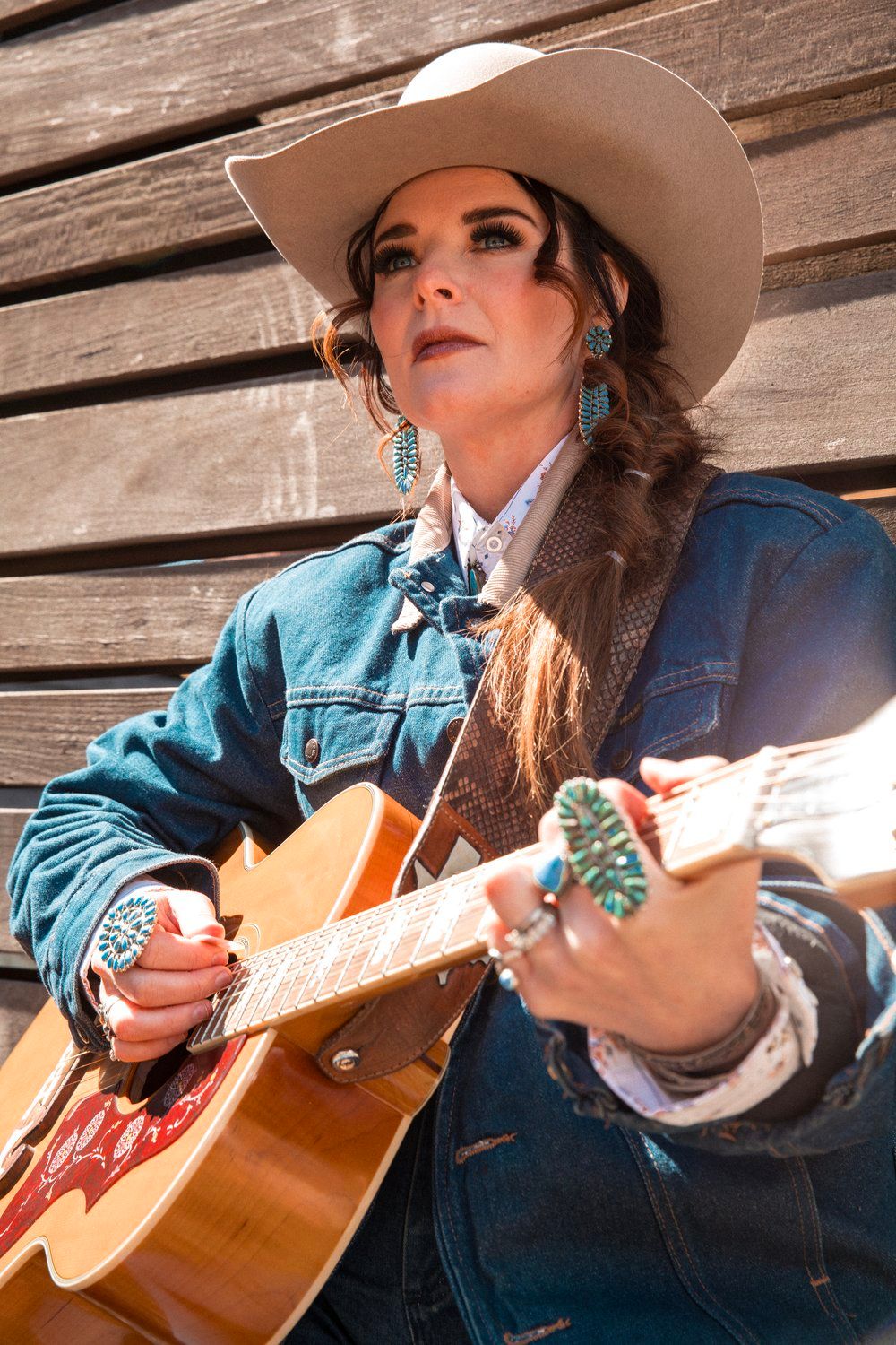 Western musician in denim jacket and cowboy hat playing acoustic guitar against wooden wall.