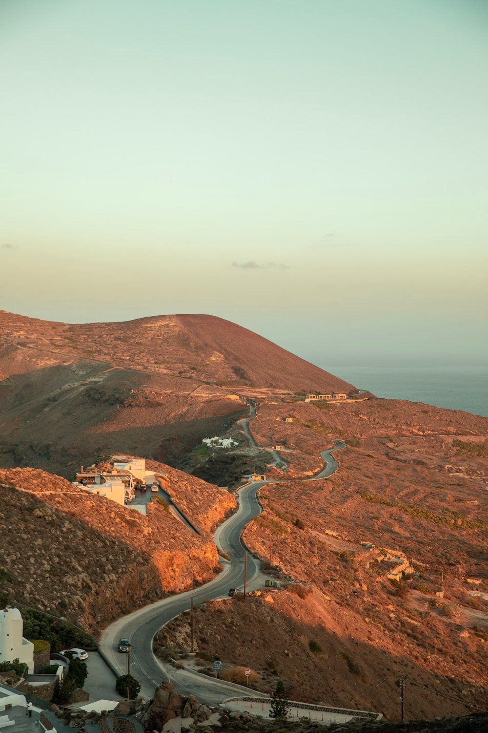 Winding mountain road through reddish terrain at sunset with coastal views.