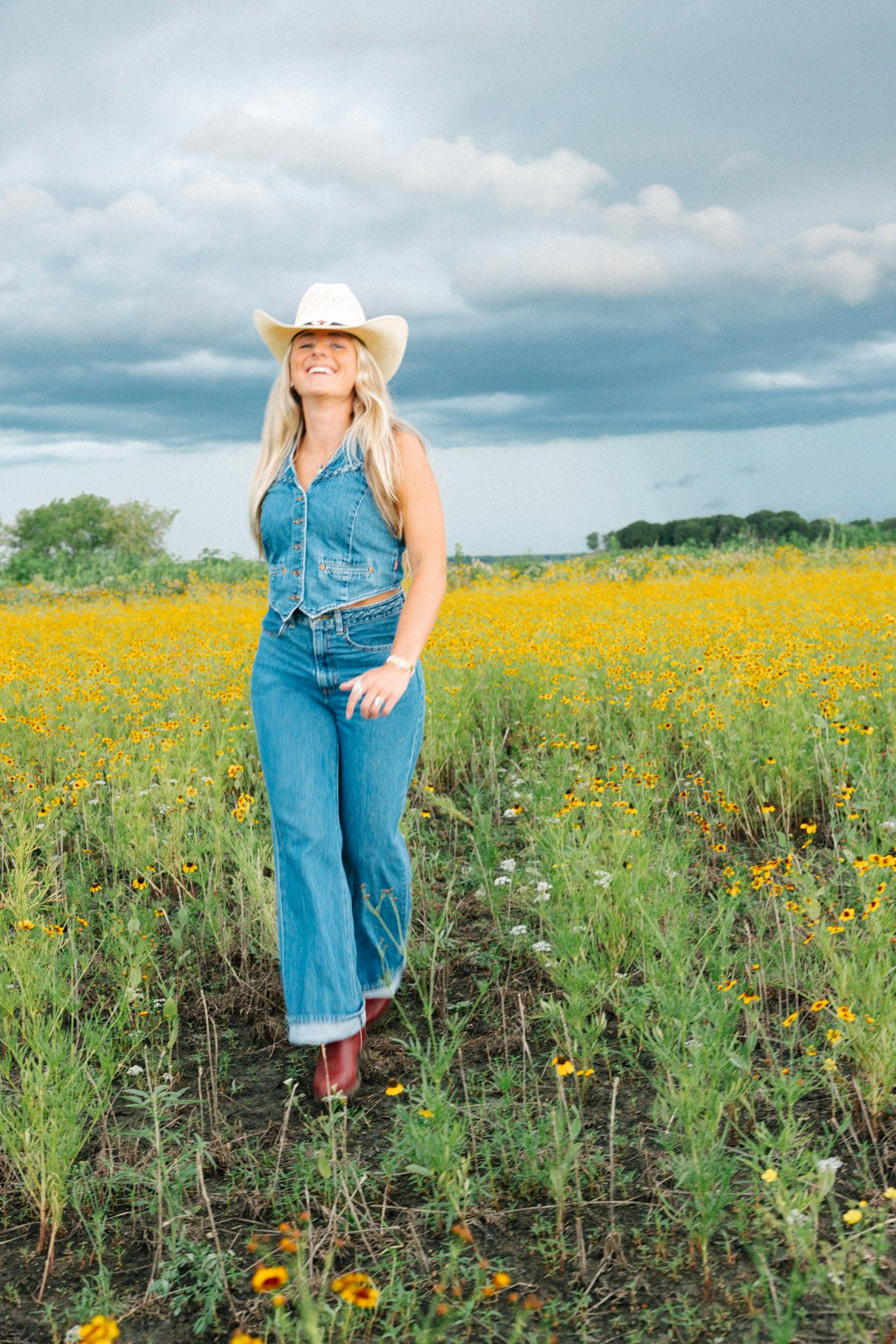 A smiling blonde woman in denim overalls and a white cowboy hat stands in a vibrant yellow wildflower field under cloudy skies.