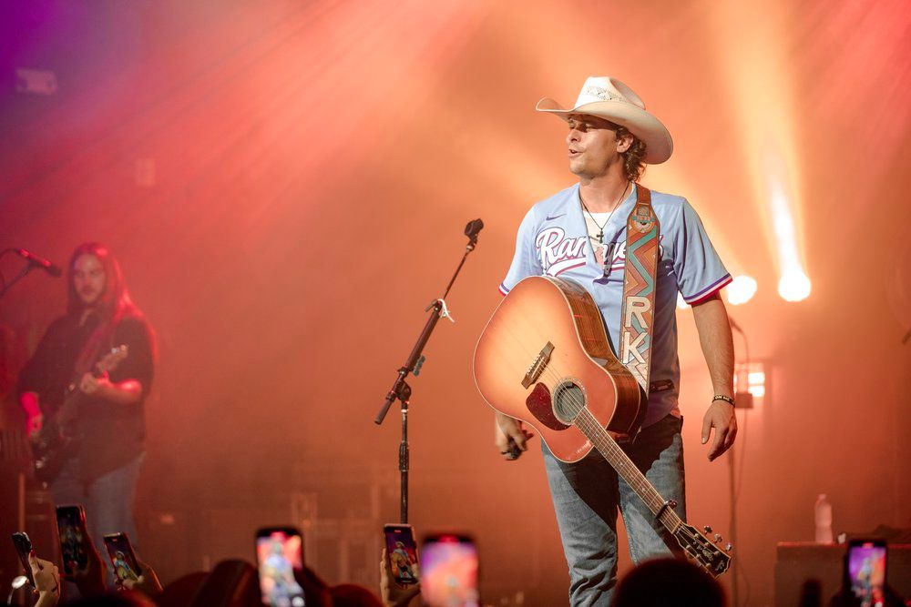Country performer Randall King in casual attire and cowboy hat plays acoustic guitar under warm stage lights.