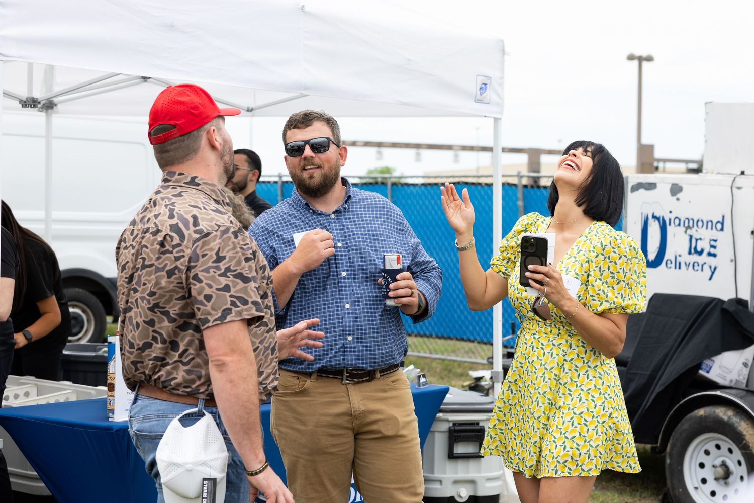 Three people socializing outdoors at a festival event, laughing and holding drinks near a white tent.