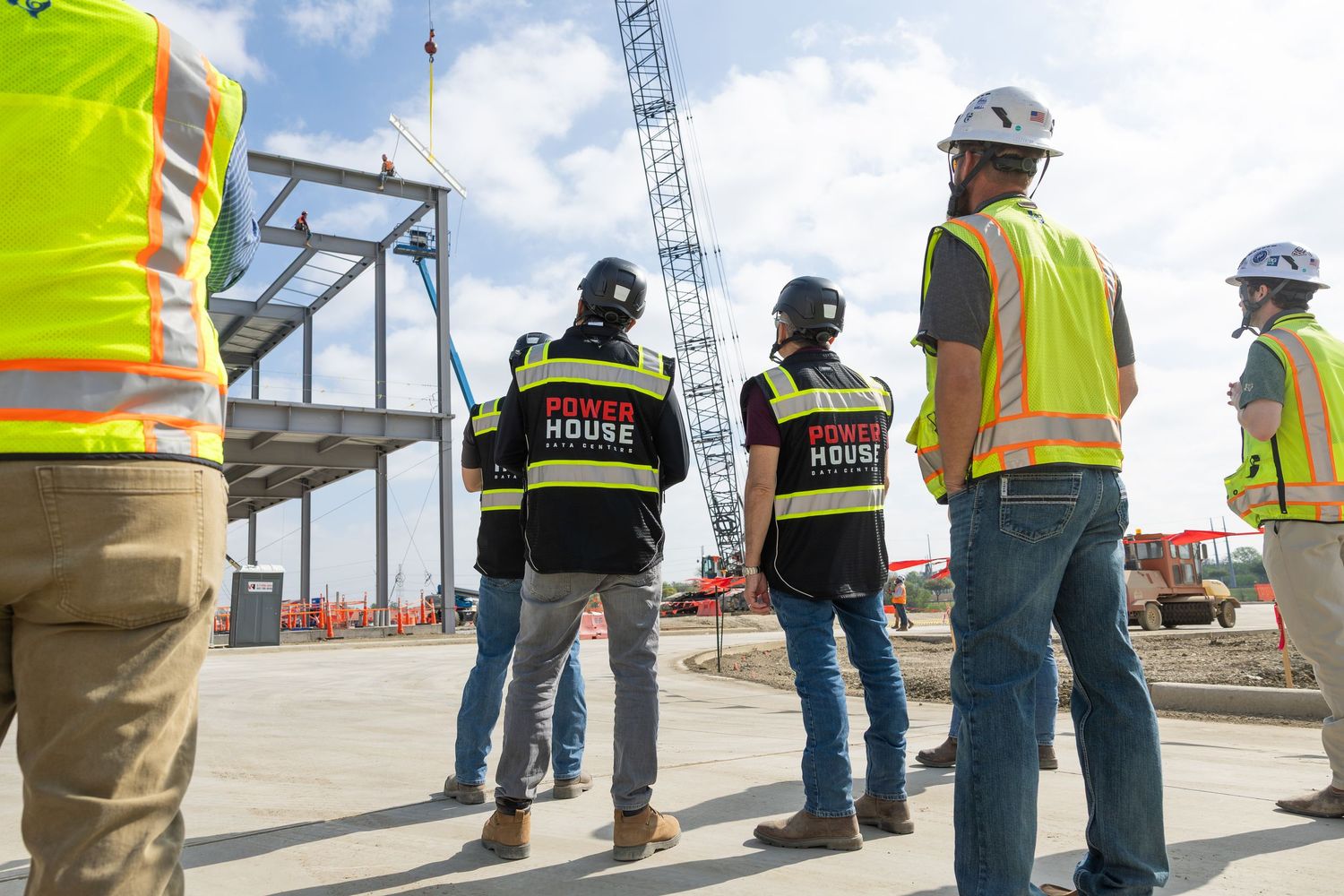 Construction workers in safety vests and Power House shirts survey a building site with steel framework and crane.