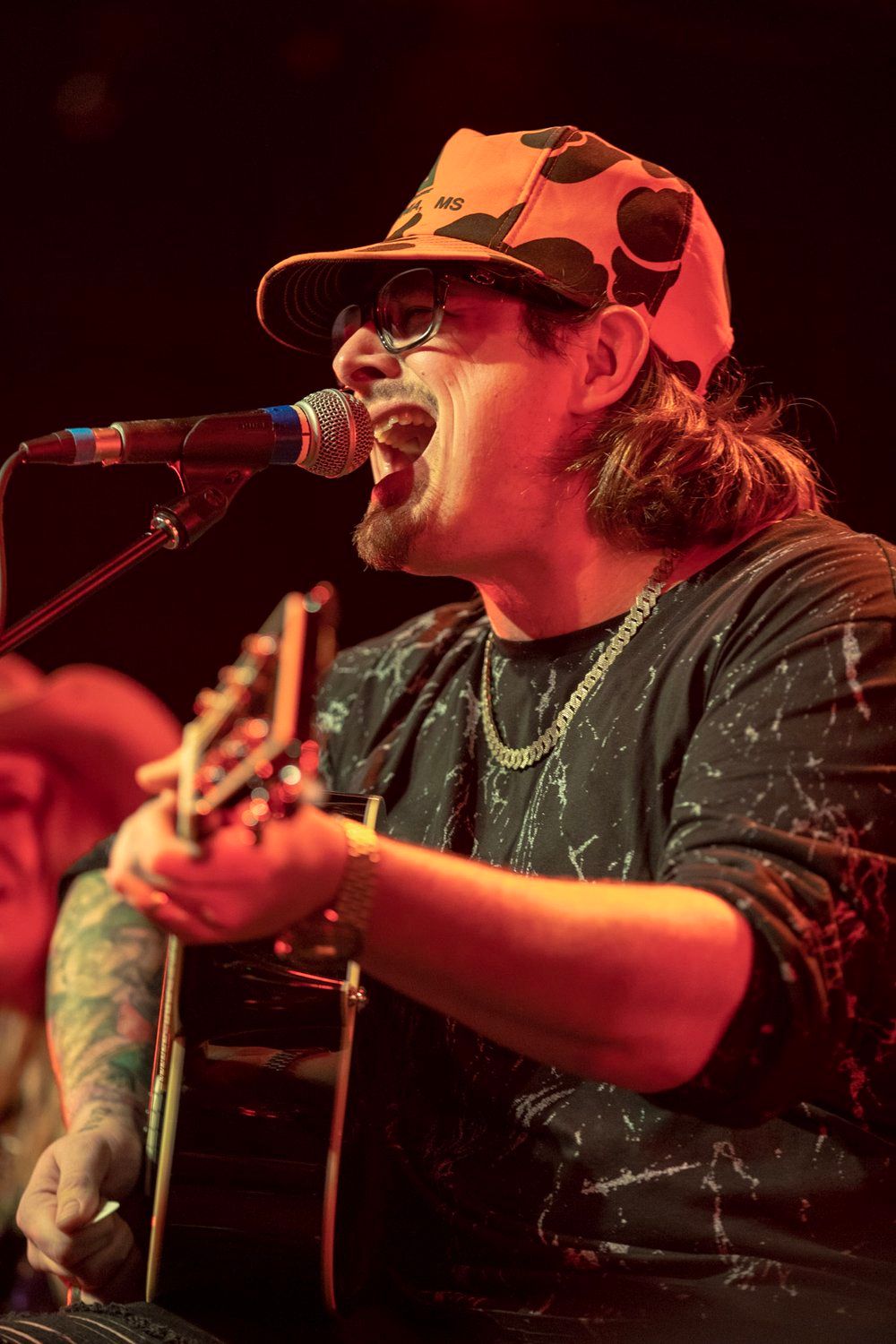 Musician Hardy wearing baseball cap performs passionately while playing acoustic guitar under red stage lighting.