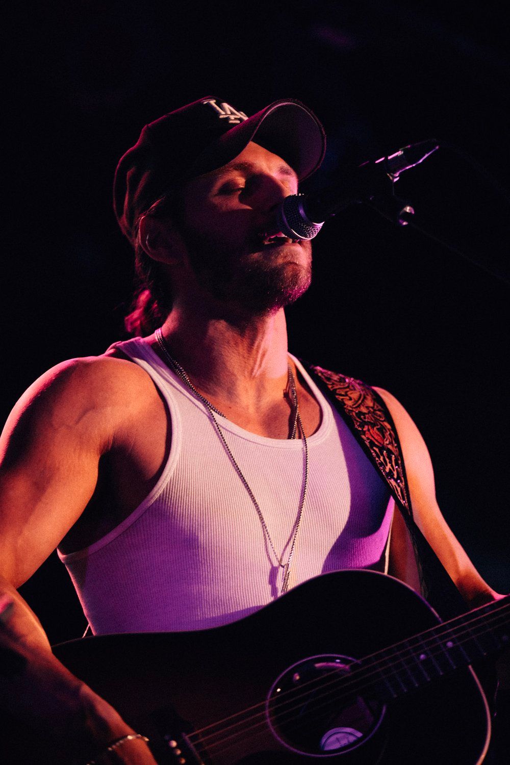 A performer Warren Zeiders in a white tank top and baseball cap holds an acoustic guitar on stage under dramatic purple lighting.